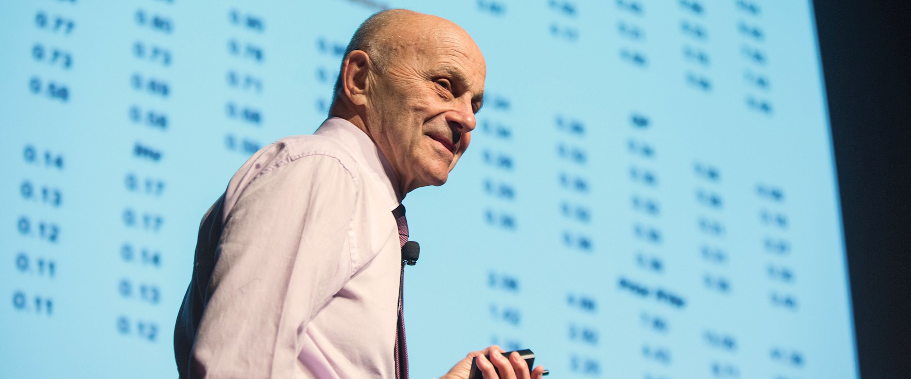 Eugene Fama presenting in front of a projector screen full on numbers